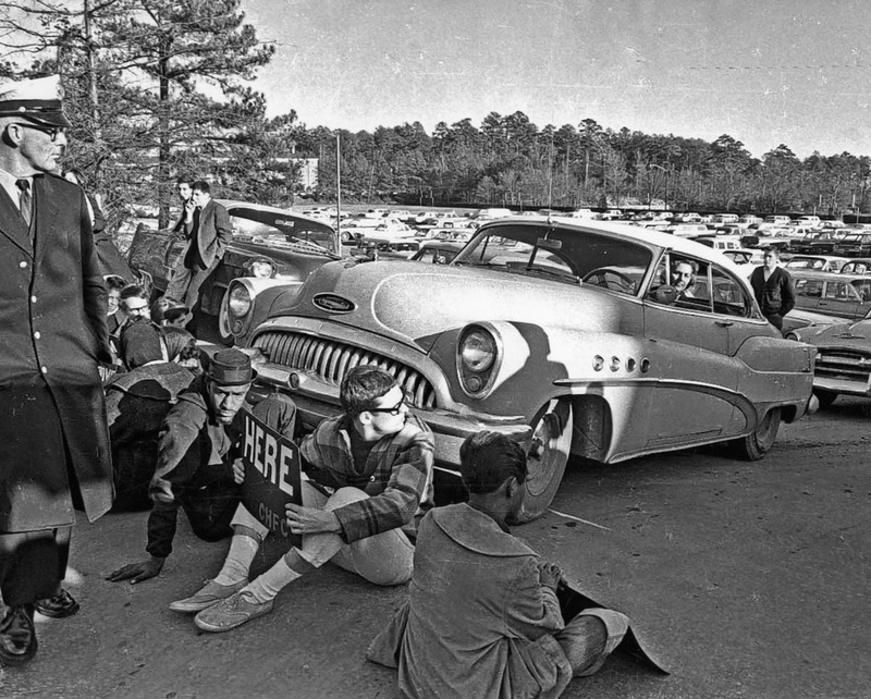 A woman stares at protesters who block her car from exiting the university parking lot.