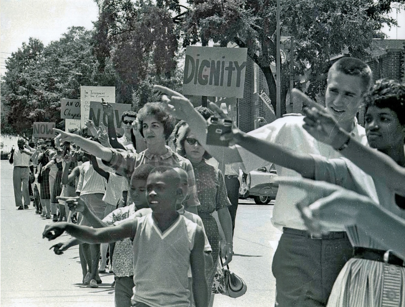 Young student marchers, both Black and white, point accusingly at segregated businesses in Chapel Hill.